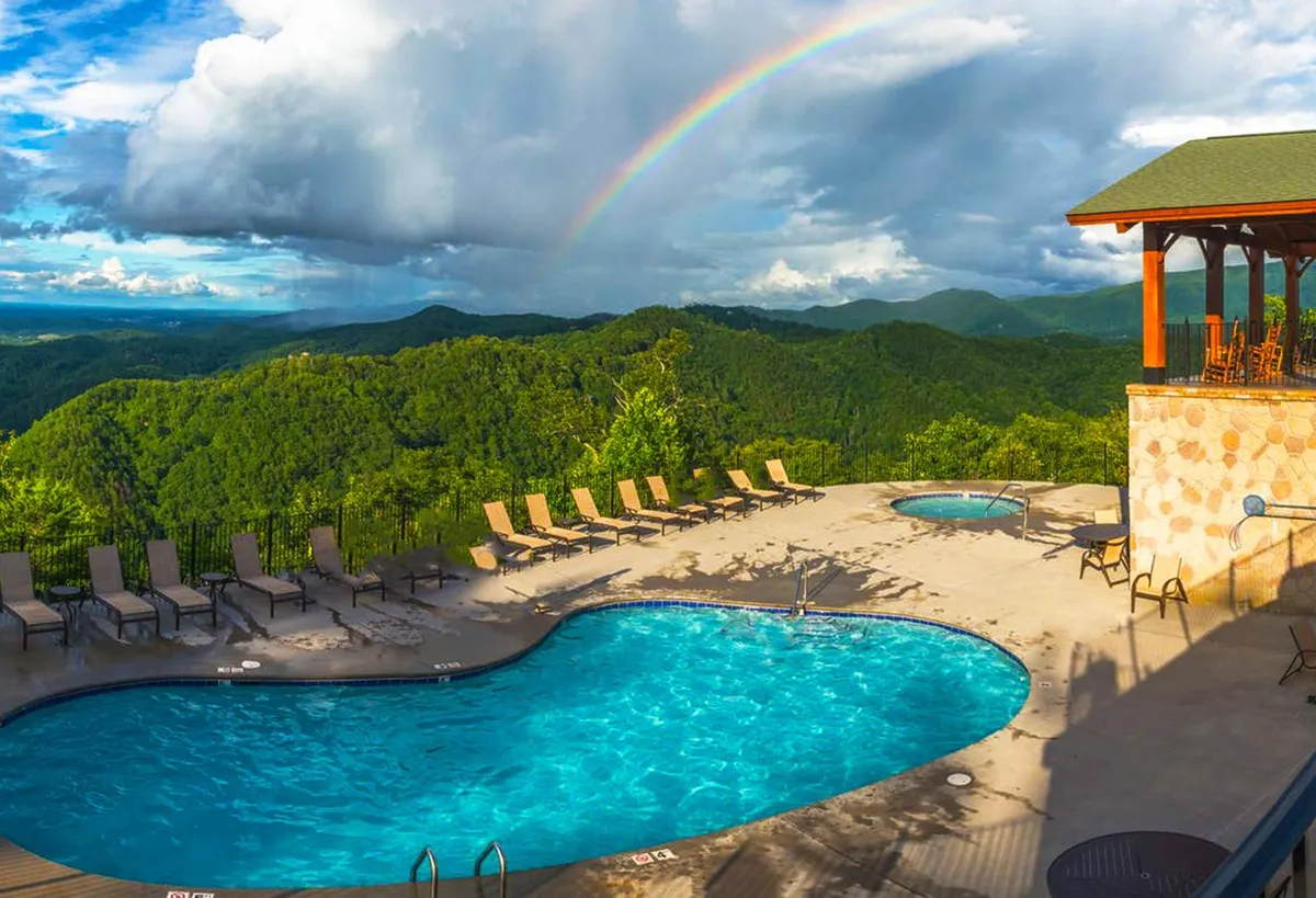This exterior image showcases a sparkling swimming pool surrounded by inviting lounge chairs and hot tubs, set against a breathtaking backdrop of the Smoky Mountains. A faint rainbow is visible in the sky, enhancing the tranquil and uplifting mood. Guests can be seen swimming, sunbathing, and socializing, all while enjoying panoramic mountain views.