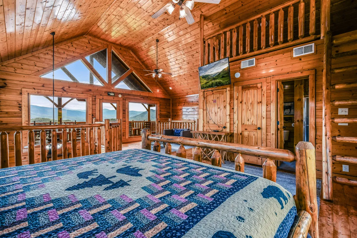 A king-size log cabin bed is the focal point of a warmly lit bedroom, framed by large windows offering sweeping mountain views. Natural wood walls and ceiling, a quilted bedspread, and a wall-mounted TV contribute to the space's rustic luxury and comfort. The ambiance radiates tranquility and invites deep relaxation after a day of adventure.