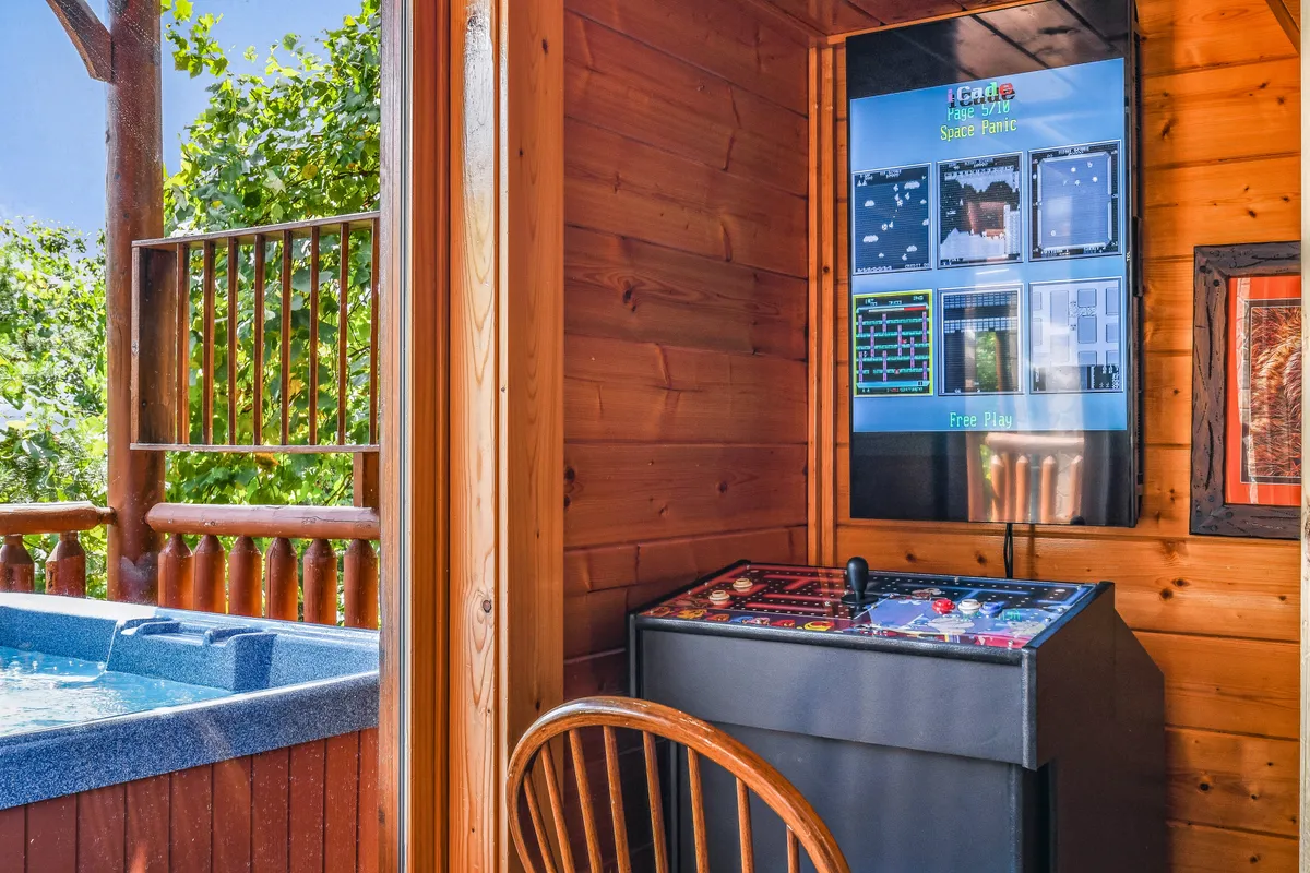 An open loft area featuring playful elements: a hot tub, arcade game setup, and classic wooden walls. The scene evokes a sense of fun and adventure, perfect for younger guests or the young at heart seeking a camp-like escape.