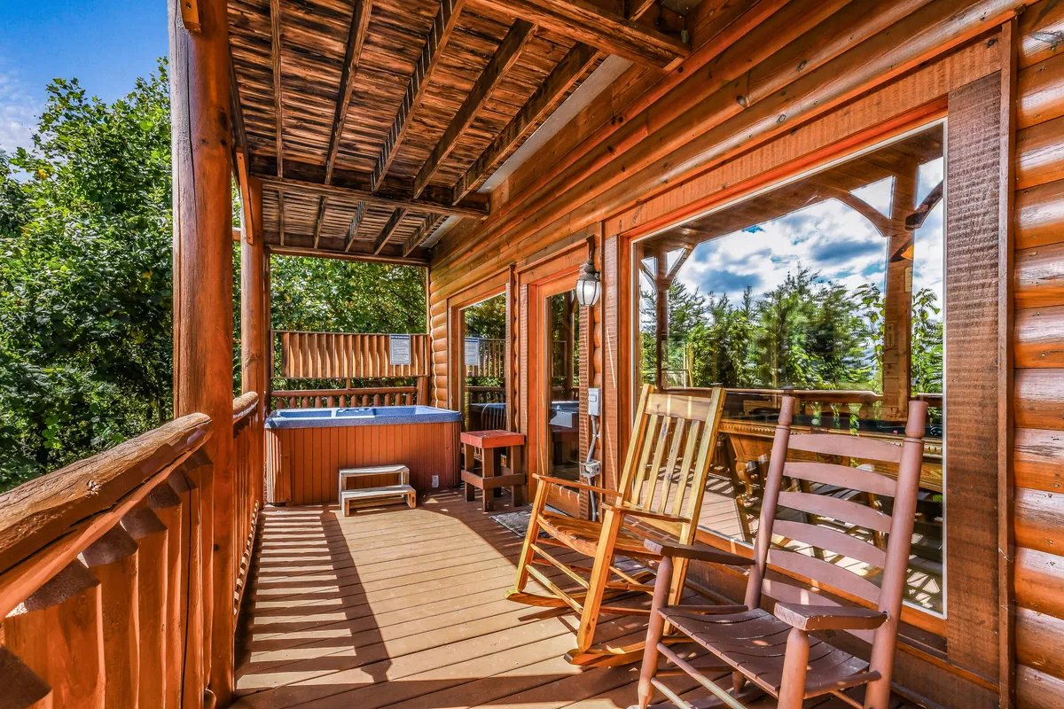 An outdoor deck with rocking chairs and a relaxing hot tub overlooks the verdant surroundings. The spacious wooden railing and large cabin windows complement the tranquil setting, suggesting a perfect place to unwind after exploring local attractions.