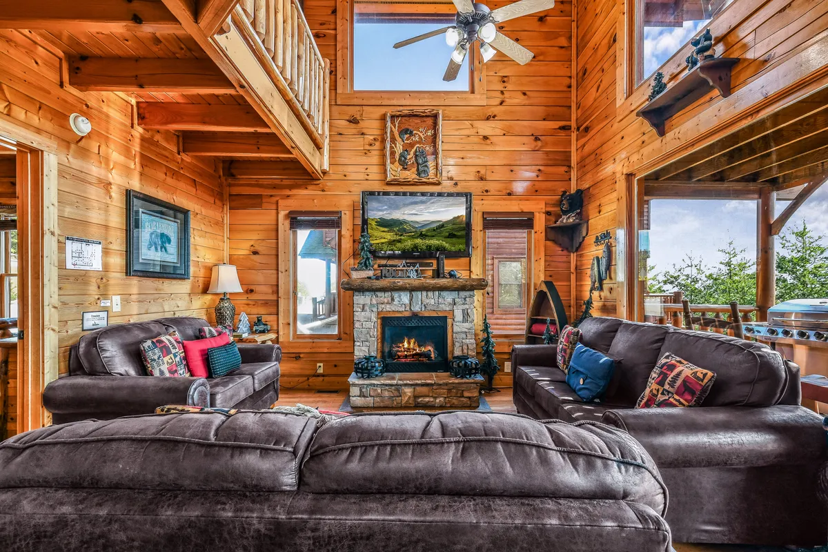 A cozy living room featuring a prominent stone fireplace framed by wooden beams, plush leather sofas arranged for conversation, expansive floor-to-ceiling windows that showcase breathtaking mountain views, and decorative artwork bringing warmth to the space.
