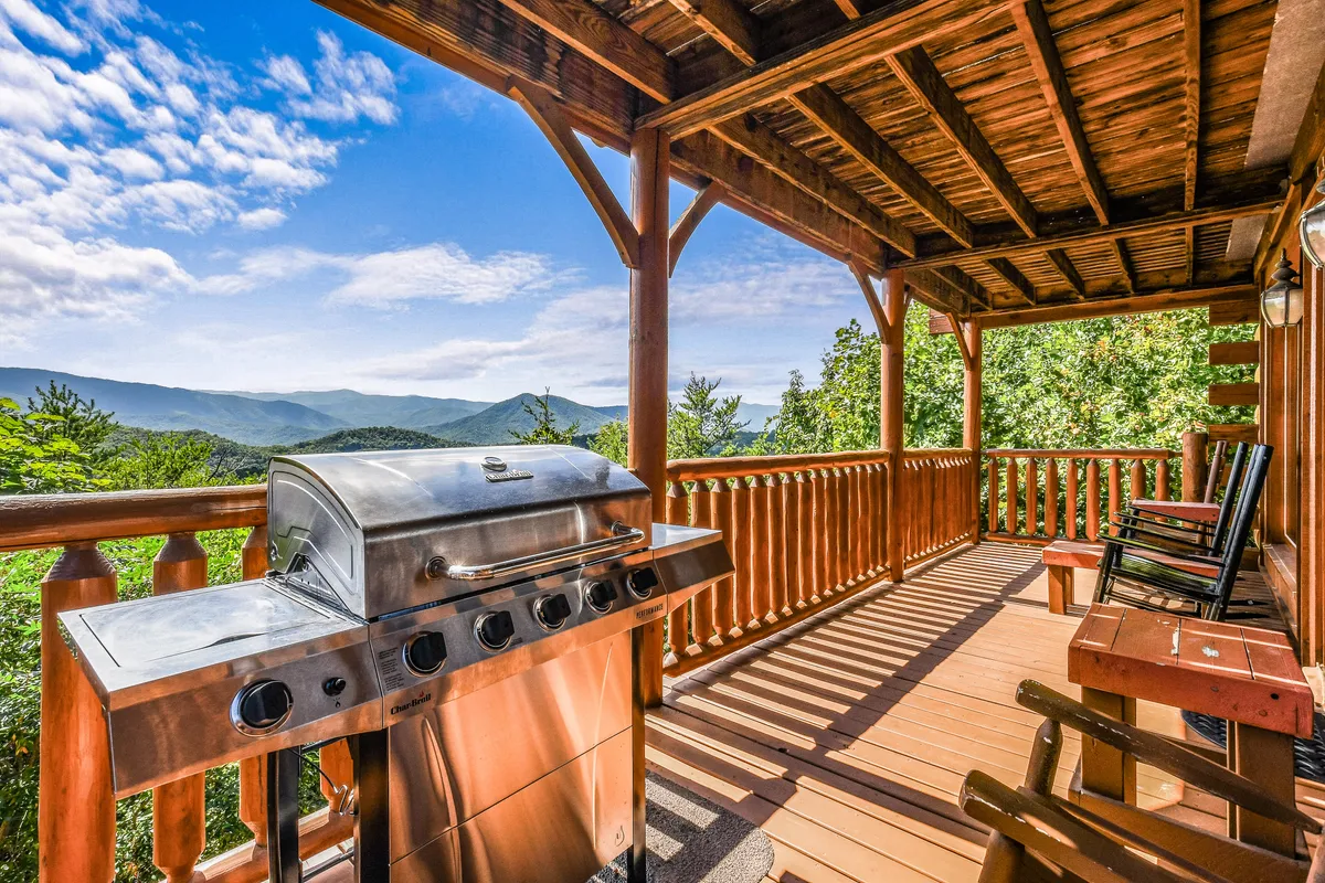 A spacious deck featuring a grill, wooden railing, and comfortable rocking chairs, all oriented toward a stunning mountain view. The scene is sunlit and embodies both comfort and the grandeur of the landscape.