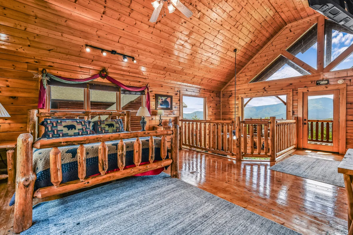 This inviting exterior shot features a cozy log bed inside a cabin bedroom, framed by large windows with views of the surrounding landscape and rustic wooden railings. The natural light streaming in hints at the scenic beauty just outside the cabin, creating a seamless connection between indoor comfort and outdoor adventure.