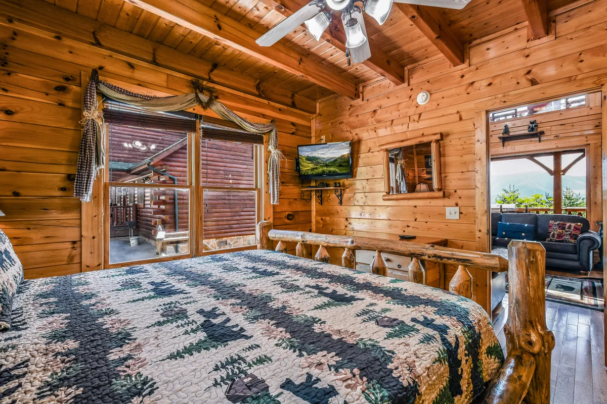 A tranquil king bedroom furnished with a log bed, quilted bedspread, wooden beams overhead, and a TV mounted on the wall. Large windows frame scenic mountain vistas, and the decor blends rustic charm with modern comfort.
