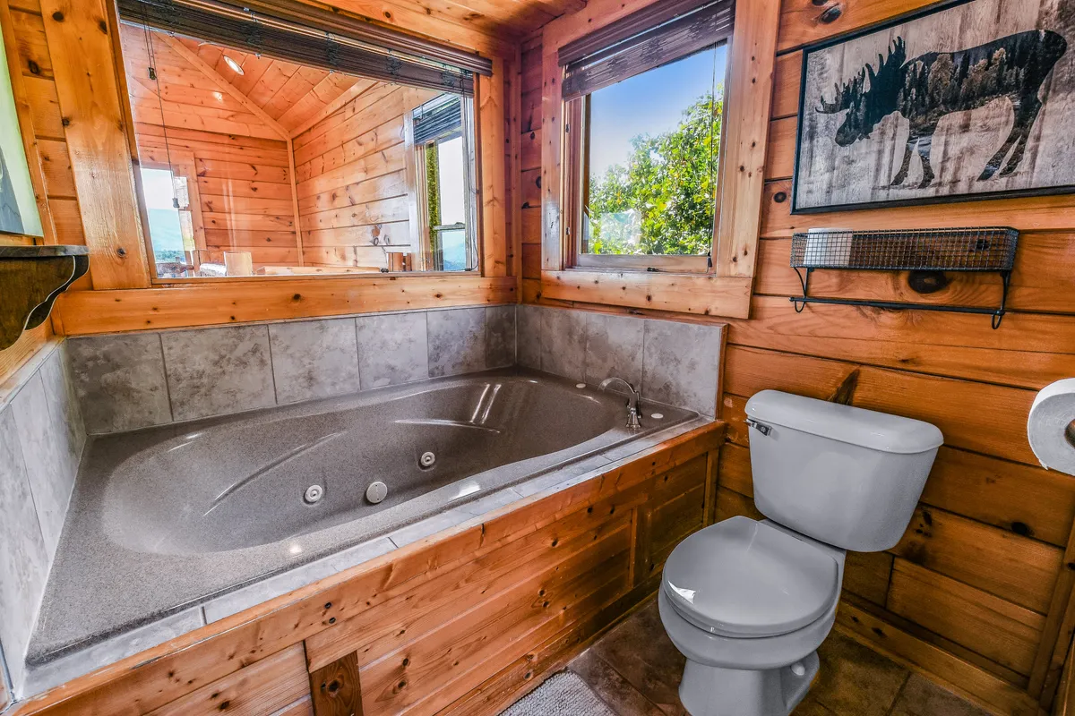 A sunlit bathroom featuring a spacious Jacuzzi tub set against wooden paneling, with large windows that bring in natural light and views. Moose artwork adds a whimsical, cabin-like touch, and the tub offers a serene spot for relaxation.